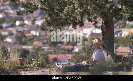 Der Mann mit dem Hut und einem Laptop sitzt auf dem Gras unter dem Baum und genießt die Aussicht Stockfoto