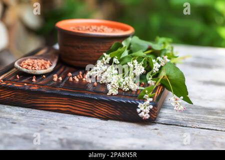 Buchweizensamen-Nahaufnahme. Geschältes Futtergetreide in einem Haufen auf einem alten Holztisch. Stillleben mit einem Strauß Buchweizenblüten in der Sommerküche. Stockfoto