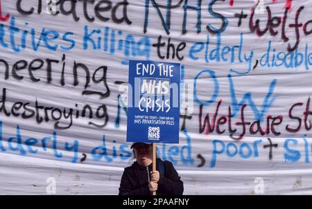 London, Großbritannien. 11. März 2023. Ein Protestteilnehmer hält ein Schild mit dem Titel "Ende der NHS-Krise" vor einem großen Banner, auf dem steht, dass die Konservativen den NHS während der Demonstration in der Tottenham Court Road zerstören. Tausende von Menschen marschierten durch das Zentrum von London, um den NHS (National Health Service) und NHS-Arbeiter zu unterstützen und gegen die Privatisierung des NHS zu protestieren. Kredit: SOPA Images Limited/Alamy Live News Stockfoto