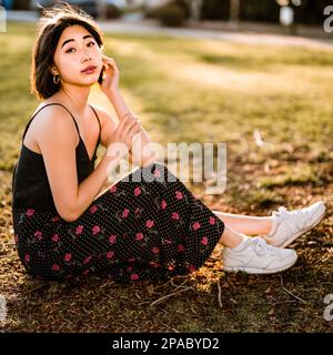 Wunderschöne junge Asiatin sitzt auf dem Gras in einem Park bei Sonnenuntergang | Seitenansicht | Blickrichtung Kamera | Hintergrundbeleuchtung Stockfoto