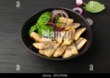 Gesalzenes Heringsfilet in der Schüssel, eingelegtes rohes Fischfleisch mit Kräutern und Gewürzen, marinierter Hering auf schwarzem Steinhintergrund Stockfoto