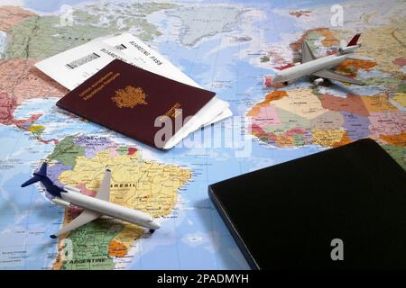 Weltkarte mit darauf, zwei Spielzeugflugzeugen, einem geschlossenen schwarzen Notizblock und einem französischen Pass mit 2 Flugtickets im Inneren. Stockfoto