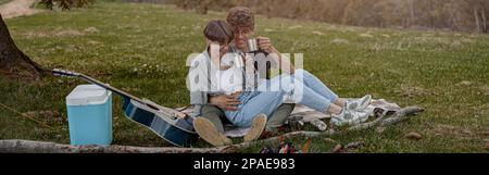 Fröhliche junge Touristen, die in Umarmung sitzen und beim Picknick lachen. Wochenende. Stockfoto