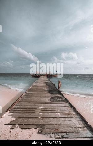 Eine vertikale Aufnahme eines sehr langen hölzernen Pier auf einer maledivischen Strandinsel, die zu einer einfachen, aber wunderschönen Gondelfahrt führt, die im grauen Meerwasser vor Anker liegt Stockfoto