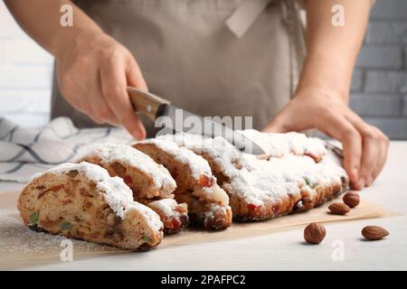 Eine Frau schneidet traditionelle Weihnachtsstollen mit Puderzucker auf einem weißen Holztisch, Nahaufnahme Stockfoto