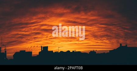 Panoramablick auf einen hellen orangefarbenen Sonnenuntergang, der die Silhouetten der Gebäude und Häuser der Stadt kontrastiert; das staubige Szenario umgibt die Stadt in einem Al Stockfoto