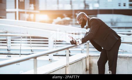 Ein ausgefallener, kahler, schwarzer Afro-Mann in einem maßgeschneiderten schwarzen Anzug, einer Brille und einer Krawatte, die sich am Geländer anlehnt, um den Papierkram besser lesen zu können Stockfoto