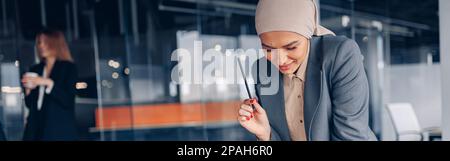 Lächelnde muslimische Geschäftsfrau im Hijab, die mit Dokumenten arbeitet und Seite anschaut Stockfoto