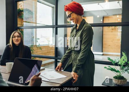Fröhliche muslimische Geschäftsfrau, die sich mit ihren Kollegen in einem modernen Sitzungssaal trifft. Glücklicher Geschäftsmann, der ein Kopftuch in einem Multicu trägt Stockfoto