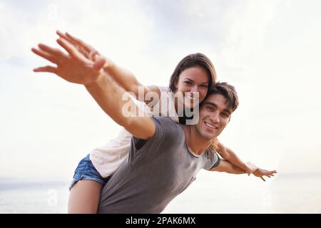 Lass uns zusammen wegfliegen. Eine kurze Aufnahme eines jungen Mannes, der seiner Freundin eine Huckepackfahrt am Strand gibt. Stockfoto