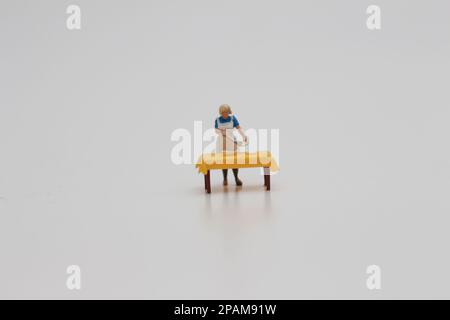 Nahaufnahme einer Miniaturfigur einer Frau, die Essen auf einem Tisch serviert, isoliert auf weißem Hintergrund. Miniaturfigur Fotokonzept. Stockfoto