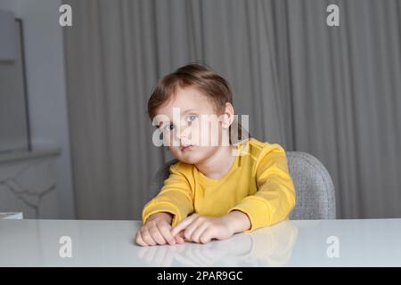 Ein nachdenklicher Junge mit braunem Haar im gelben Pullover, der am Tisch sitzt, in die Kamera schaut und an den Händen zieht Stockfoto