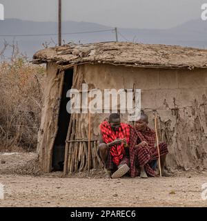 Karatu, Tansania - 16. Oktober 2022: Zwei masai-Männer sitzen neben einer Hütte in ihrem Dorf mit kleinen Schlammhütten, umgeben von einem Dornzaun. Stockfoto