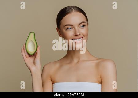 Beauty-Hautpflegekonzept. Positiv gesund zufriedene Frau sieht zärtlich beiseite Lächeln behutsam hält die Hälfte des Avocados hat perfektes Make-up weiße Zähne Wrapp Stockfoto