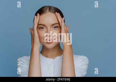 Charmantes junges braunes weibliches Model berührt das Gesicht genießt weiche Haut hat natürliches Make-up gekleidet in legerem weißen T-Shirt sieht ruhig vor der Kamera aus Stockfoto