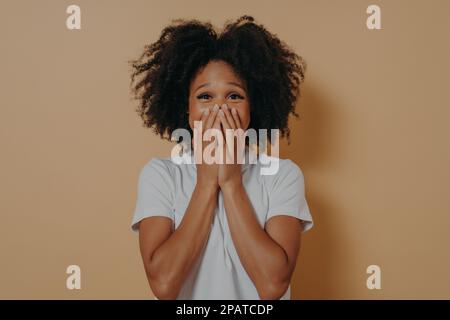 Überraschend aufgeregte, dunkelhäutige Frau in weißem T-Shirt, das ihren Mund bedeckt, mit isolierten Händen auf pastellfarbenem beigefarbenem Hintergrund mit Kopierbereich. Positive Frauen Stockfoto