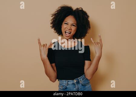 Rockstar. Junge selbstbewusste afro-amerikanische Frau, die beide Hände im Rock 'n' Roll-Schild hält und laut schreit, mit emotionalem Gesichtsausdruck, Gefühl Stockfoto