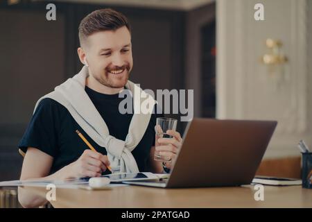 Ein glücklicher Freiberufler oder Enterpruer in zwangloser Kleidung sitzt am Schreibtisch vor dem geöffneten Laptop Computer und surft im Internet, hält ein Glas Wasser und Stockfoto