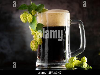 Dunkles Bier in einem Becher und grünen hop auf Holztisch auf braunem Hintergrund Stockfoto