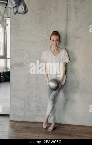 Vertikaler Schuss einer jungen, positiven Balletttänzerin in weißem T-Shirt und Leggings, die sich an die Wand lehnen und einen kleinen silbernen Fitball halten, während barr spielt Stockfoto