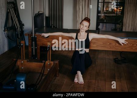 Fit junge Frau, die auf dem Boden sitzt, am Trapeztisch, der sich nach Dehnübungen oder Gymnastikübungen auf dem Pilates-Reformator ruht, sich entspannt und zufrieden fühlt Stockfoto