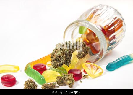Verschiedene Gummis und getrocknete medizinische Marihuana-Knospen fielen aus dem geprägten Glasgefäß. Auf weißem Hintergrund. Viel Platz Stockfoto
