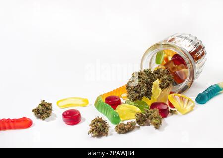 Verschiedene Gummis und getrocknete medizinische Marihuana-Knospen fielen aus dem geprägten Glasgefäß. Auf weißem Hintergrund. Viel Platz Stockfoto