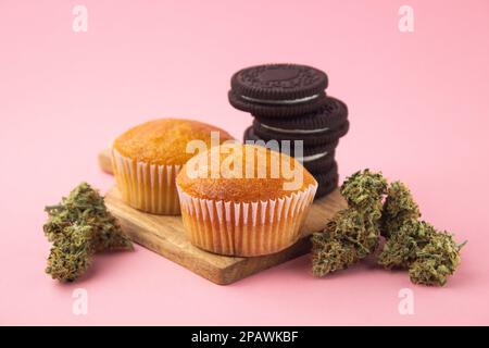 Cannabis-Cupcakes und Schokoladenkekse auf einem Holzbrett, trockene Marihuana-Knospen an den Seiten. Auf pinkfarbenem Hintergrund Stockfoto