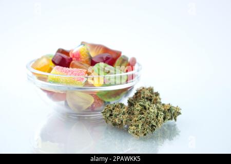 Verschiedene Gummibonbons in einer kleinen Glasschüssel, in der Nähe im Vordergrund mehrere Knospen getrockneten medizinischen Marihuanas, reflektiert auf dem Glas Stockfoto
