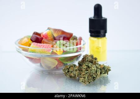 Verschiedene Gummibonbons in einer kleinen Glasschüssel und einer Flasche CBD-Canna-Öl, in der Nähe im Vordergrund mehrere Knospen getrockneten medizinischen Marihuanas, Stockfoto