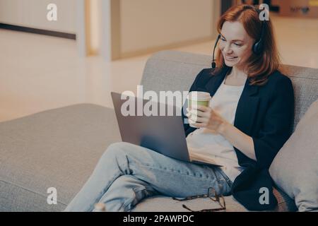 Lächelnde junge Callcenter-Agentin mit schnurlosem Headset und Laptop, kommuniziert mit Kunden des Finanzunternehmens, bietet Online-Beratung, Stockfoto