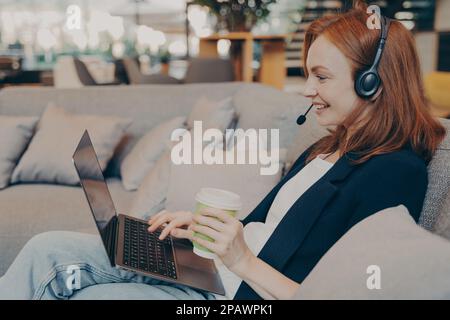 Lächelnde junge Lehrerin mit kabellosen Kopfhörern und Laptop, die Videokonferenz führt, mit Schülern per Webcam spricht, online unterrichtet, w Stockfoto