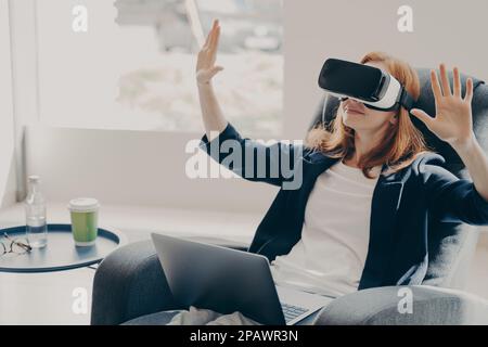 Geschäftsfrau in einer Virtual-Reality-Brille, die eine VR-Brille verwendet, innovative Geschäftsmethoden testet, auf einem Sessel mit einem geöffneten Laptop sitzt, interagiert Stockfoto