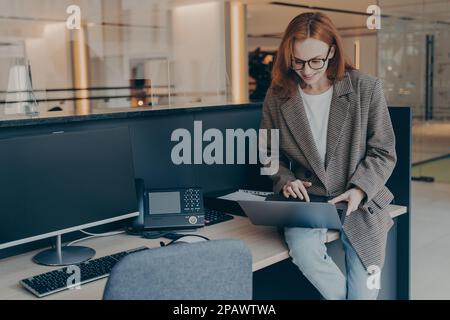 Vielbeschäftigte Frau in lässiger Kleidung, die ihren Laptop benutzt, auf dem Schreibtisch sitzt, im Internet surft und recherchiert mit dem Computer, rothaarige Frau, die in Mod arbeitet Stockfoto