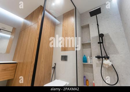 Badezimmer in modernem Stil mit grauen und weißen Fliesen. Es gibt einen großen Spiegel mit leuchtenden Lampen, Tischplatte mit Holzschubladen und Waschbecken, Bad mit Stockfoto