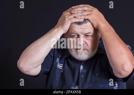 Schönes Porträt eines ukrainischen, bärtigen Seniorenmannes, der über Lebensprobleme nachdenkt, während er in der Dunkelheit sitzt Stockfoto