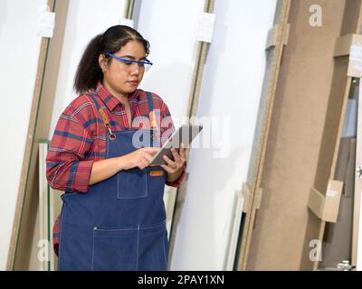 Eine Frau mittleren Alters, die ihre E-Mail-Bestellung auf einem Tablet-Computer überprüft. Das Sperrholzlagerfach befindet sich im Hintergrund. Asiatischer Zimmermann arbeitet in einem Ofen Stockfoto