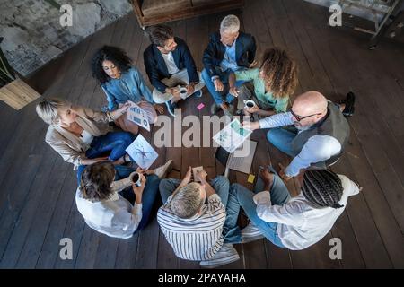 Eine vielfältige Gruppe von Fachleuten nimmt an einem kreativen Brainstorming Teil, setzt sich auf den Boden und teilt Ideen. Mit Laptops, Tablets und Notizbüchern Stockfoto