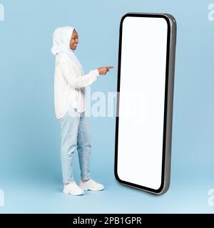 Die ganze Körperlänge einer schwarzen muslimischen Frau, die ein großes Smartphone mit leerem weißen Bildschirm benutzt und auf das Modell zeigt Stockfoto