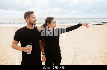 Glückliches junges Sportpaar, das sich nach dem Joggen oder Workout am Strand ausruht und eine Dame, die wegzeigte Stockfoto