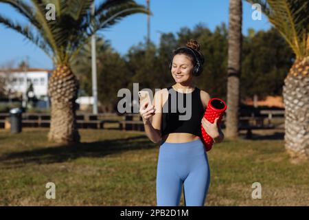 Lächelnde junge Frau macht Pause beim Training, steht mit rotem Massagerollzylinder, benutzt Smartphone, hört Lieder in Kopfhörern, trägt Sportbekleidung Stockfoto