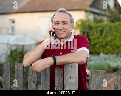 Ein erwachsener Mann, der entspannt neben dem Zaun steht und im Garten telefoniert und lächelt Stockfoto