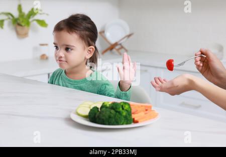 Mutter füttert ihre Tochter in der Küche, Nahaufnahme. Ein kleines Mädchen, das sich weigert, Gemüse zu essen Stockfoto