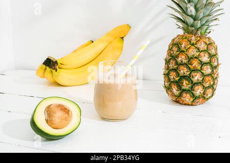 Bananen-, Ananas- und Avocado-Smoothie auf einem weißen Holztisch bei Sonnenlicht. Minimal-Entgiftungsdiät-Konzept, Sommer-Vitamingetränk. Stockfoto