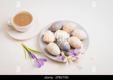 Grauer Teller mit lackierten Ostereiern, Kaffeetasse und Krokusblumen auf weißem Hintergrund. Osterkonzept. Draufsicht. Stockfoto