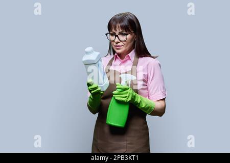 Frau mittleren Alters in Handschuhschürze mit organischen Reinigungsmitteln auf grauem Hintergrund Stockfoto