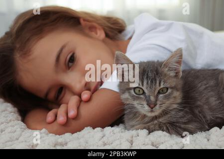 Süßes kleines Mädchen mit Kätzchen auf weißer Decke drinnen, Nahaufnahme. Haustier aus der Kindheit Stockfoto