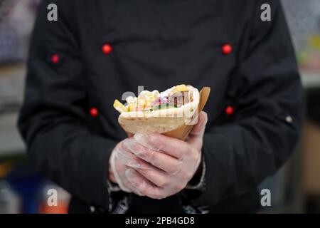 Der Koch hält Gyros in Pita-Brot verpackt. Traditionelles griechisches Fast-Food-Gericht mit natürlichen Zutaten - gegrilltes Hühnerfleisch, Pommes Frites und weißer Zatz Stockfoto