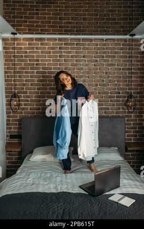 Eine schöne Frau in Designerkleidung steht auf dem Bett und hält online gekaufte Kleidung in ihren Händen. Vertikaler Rahmen. Stockfoto