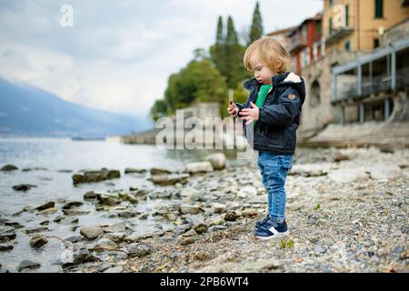 Süßer kleiner Junge, der im Hafen von Varenna, einer der malerischsten Städte am Ufer des Comer Sees, mit Kieselsteinen spielt. Varenna, Lombardei, Italien. Stockfoto